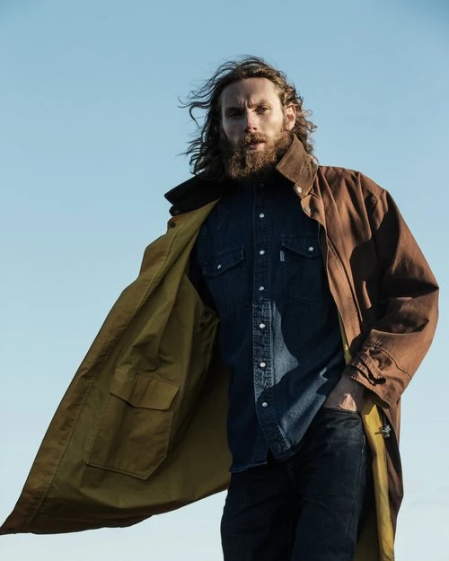 A man with long hair and a beard stands outdoors in a blue denim shirt, dark jeans, and an open brown coat against a blue sky.