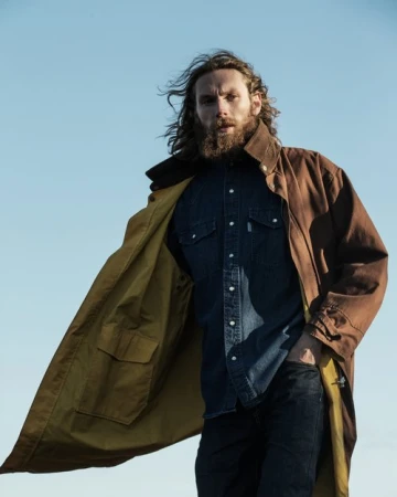 A man with long hair and a beard stands outdoors in a blue denim shirt, dark jeans, and an open brown coat against a blue sky.