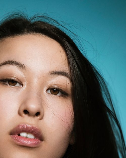 Close-up portrait of an Asian woman with brown eyes and dark hair against a vibrant teal background, showcasing beauty and fashion.