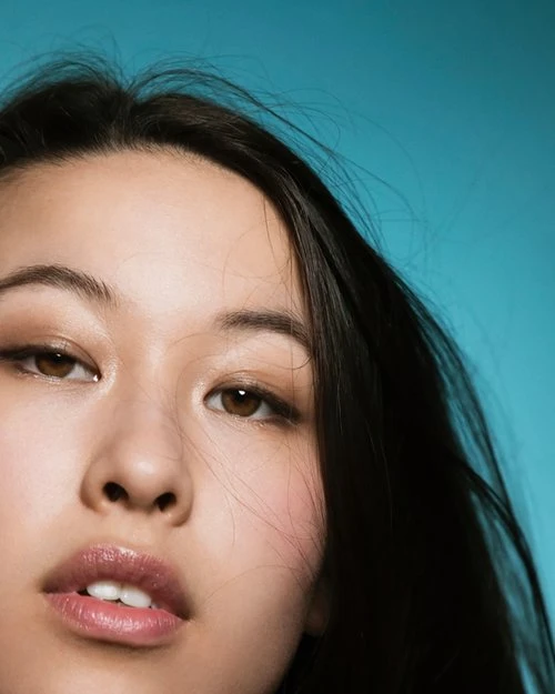 Close-up portrait of an Asian woman with brown eyes and dark hair against a vibrant teal background, showcasing beauty and fashion.