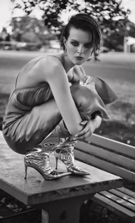 Model with dark short hair wearing a silk slip dress and shiny metallic boots crouching on a park bench