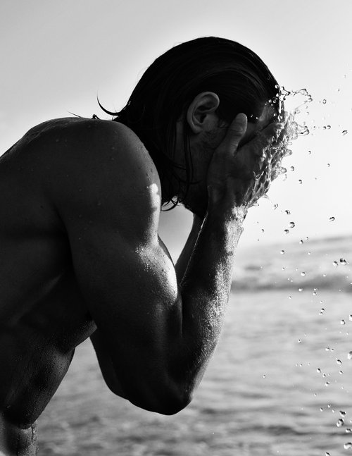 Black and white close-up of a muscular man at a beach, splashing water on his face against a bright, clear sky and rippling ocean backdrop.