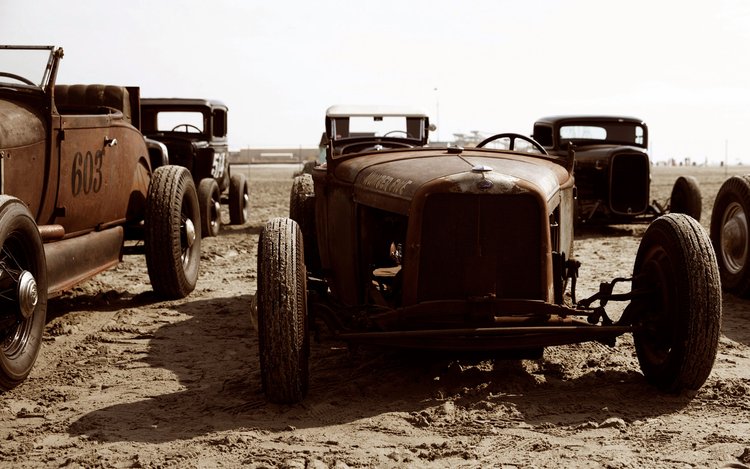 Several vintage rusty hot rod cars displayed on a beach-like setting with a sepia-toned filter applied for a retro feel.
