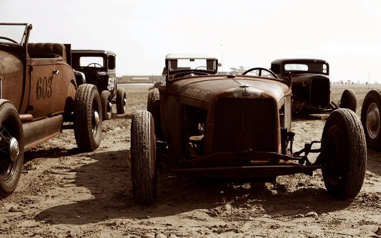Several vintage rusty hot rod cars displayed on a beach-like setting with a sepia-toned filter applied for a retro feel.