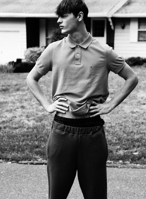 Monochrome portrait of a young man in polo shirt and sweatpants standing outside a house on a lawn with hands on his waist.