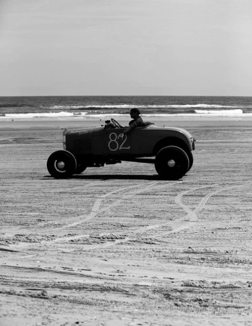 Vintage race car with driver on beach near ocean. Black and white photo shows vehicle number 82 during an automobile event by the sea.