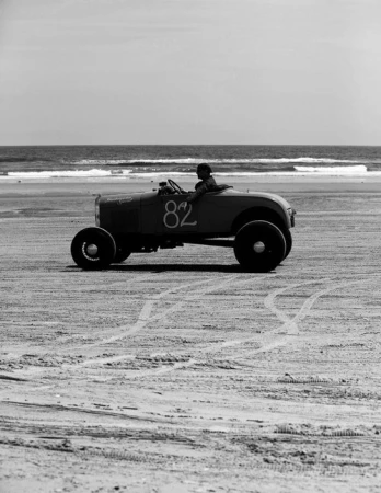 Vintage race car with driver on beach near ocean. Black and white photo shows vehicle number 82 during an automobile event by the sea.
