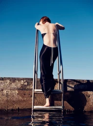 Woman in a black satin dress climbing a metal pool ladder against a clear blue sky, photographed by Todd Barry in Sydney.