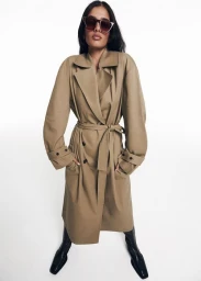 Model in dark sunglasses and a beige belted trench coat with black boots against a white seamless background