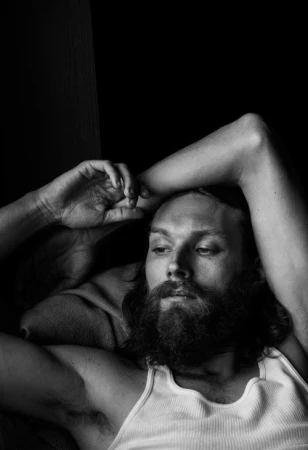 Portrait of a bearded man with long hair wearing a white tank top, resting with his arms raised against a dark background.