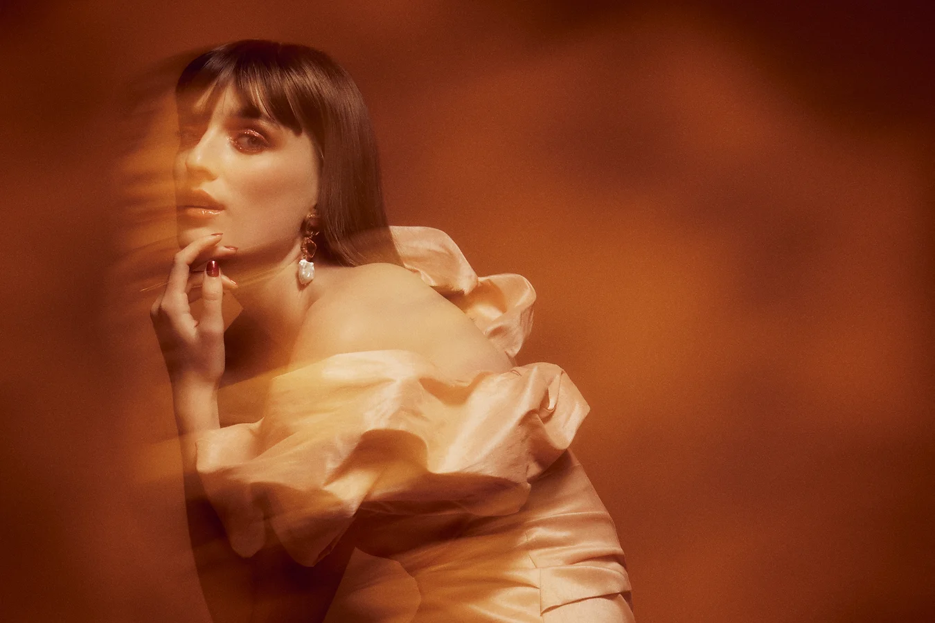 Portrait of a woman with long brown hair, wearing a light-colored dress with ruffles, posing against a blurred orange backdrop.
