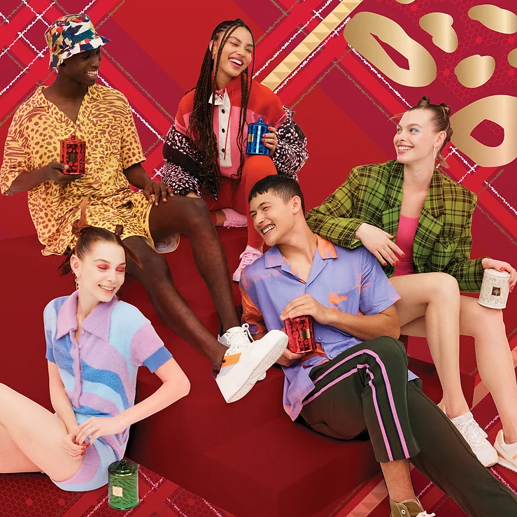 Group of five diverse young adults smiling and sitting together, holding beverage cans against a red patterned backdrop, indoors.