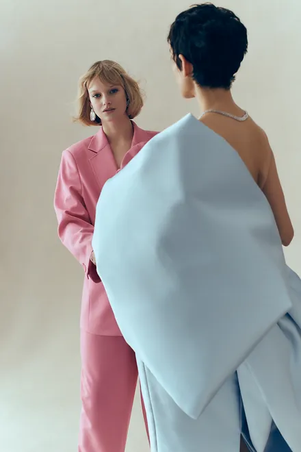 Two fashion models in a studio setting. One woman wears a pink suit, while the other displays a large light blue sculptural garment.