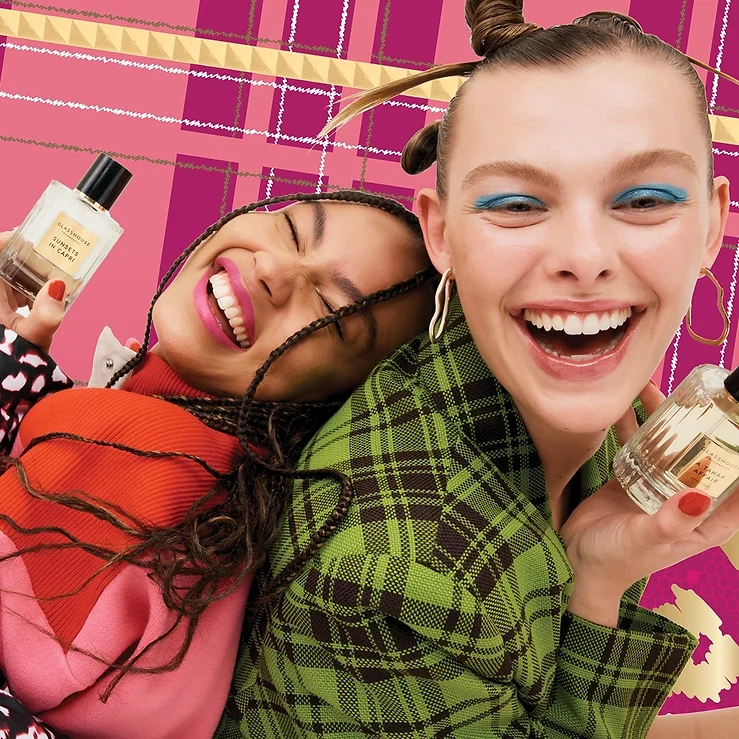 Two women smiling and holding perfume bottles in front of a pink and gold patterned background, cheerful beauty campaign.