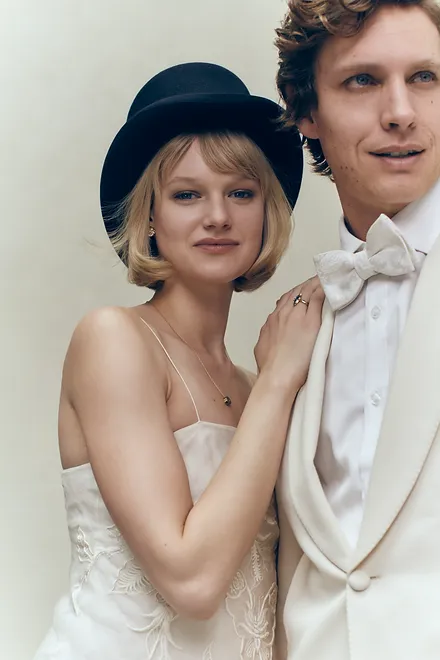 Stylish couple poses in formal attire against a neutral backdrop. The woman wears a black hat and the man sports a white bow tie.