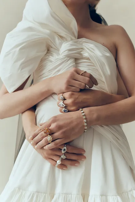 Close-up of two women embracing, hands adorned with multiple rings and a bracelet, wearing an elegant white dress in a neutral setting.