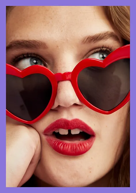 Close-up of a young woman with red lipstick and heart-shaped sunglasses, looking to the side with a slightly open mouth.