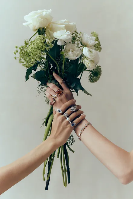 Close-up of two people holding a vibrant flower bouquet, adorned with multiple rings and a bracelet against a plain background.