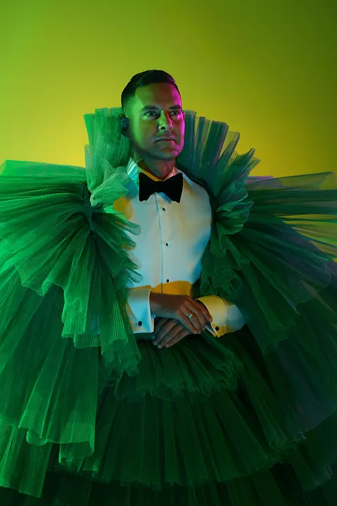 Portrait of a man in formal wear with a black bow tie and dramatic green tulle ruff and skirt, posing against a yellow-green backdrop.