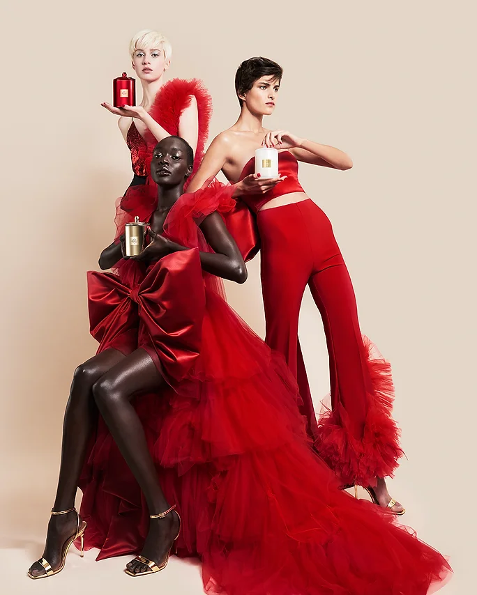 Three fashion models in red couture dresses pose in a studio while holding perfume bottles, against a neutral backdrop.