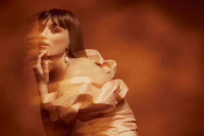 Portrait of a woman with dark hair and bangs wearing an off-shoulder peach dress against a warm brown gradient background with motion blur.