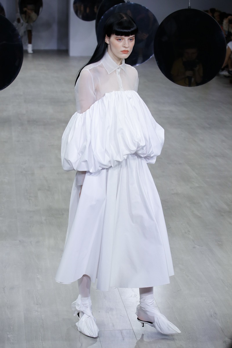 A woman walks a runway in a voluminous white dress with sheer sleeves and matching shoes during a fashion show with suspended reflective orbs.