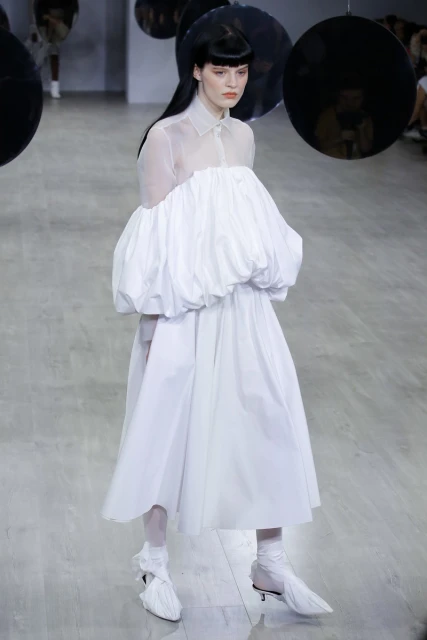A woman walks a runway in a voluminous white dress with sheer sleeves and matching shoes during a fashion show with suspended reflective orbs.