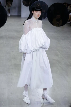 A woman walks a runway in a voluminous white dress with sheer sleeves and matching shoes during a fashion show with suspended reflective orbs.