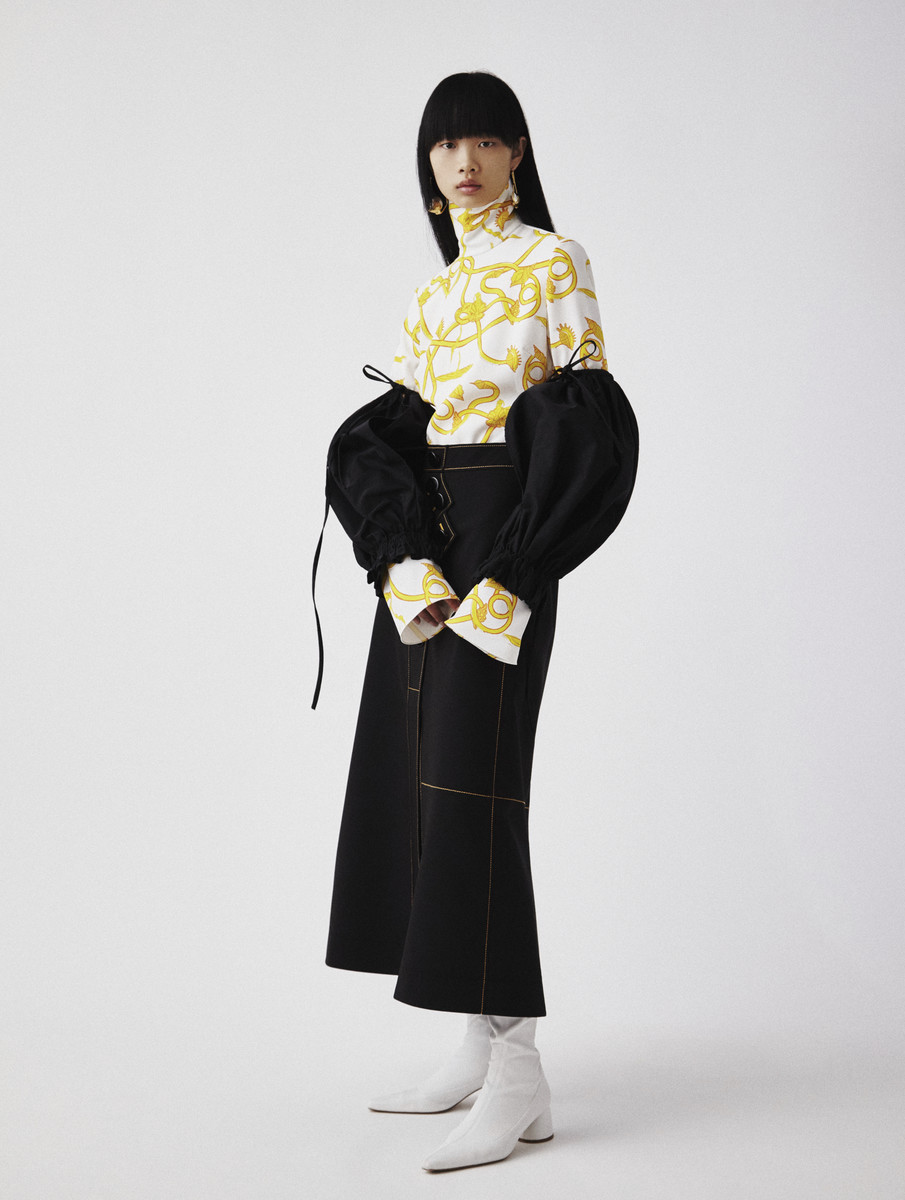 Studio shot of an Asian fashion model in a white and gold patterned top with puffy black sleeves, paired with a black skirt and white boots.
