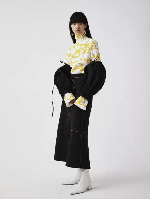 Studio shot of an Asian fashion model in a white and gold patterned top with puffy black sleeves, paired with a black skirt and white boots.
