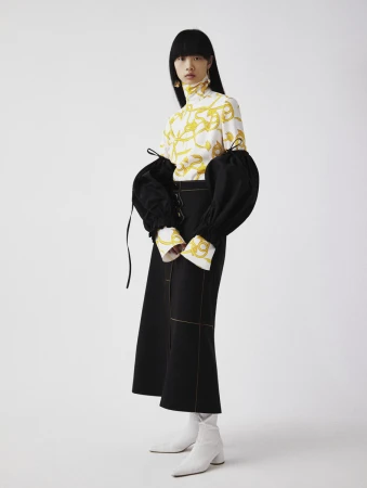 Studio shot of an Asian fashion model in a white and gold patterned top with puffy black sleeves, paired with a black skirt and white boots.