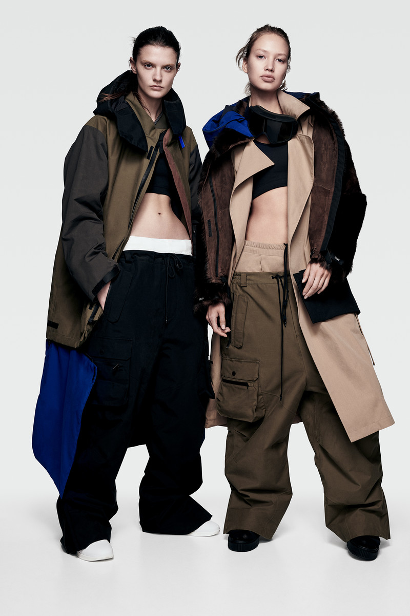 Two women showcase fashion: one in a trench coat with a fur-lined jacket, the other in a brown coat, black top, and baggy pants, against a white backdrop.