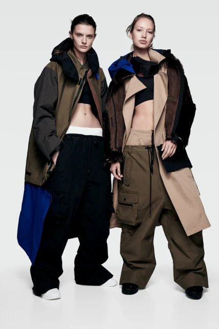 Two women showcase fashion: one in a trench coat with a fur-lined jacket, the other in a brown coat, black top, and baggy pants, against a white backdrop.