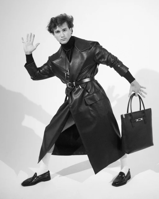 A young adult man in a black leather coat and loafers holds a black leather tote bag in a full studio shot, against a white backdrop.
