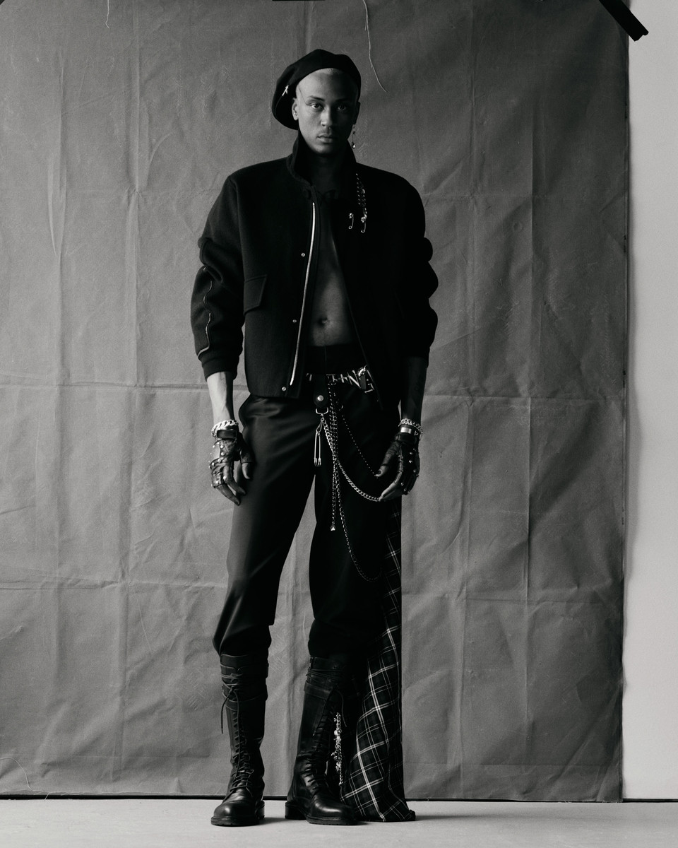 Portrait of a stylish adult male in a black beret, jacket, pants, and boots, posing indoors against a textured gray backdrop in a photo studio.