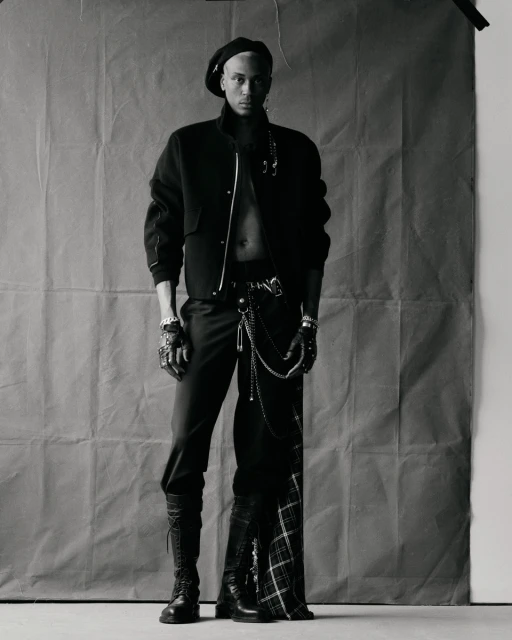 Portrait of a stylish adult male in a black beret, jacket, pants, and boots, posing indoors against a textured gray backdrop in a photo studio.