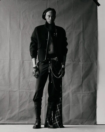 Portrait of a stylish adult male in a black beret, jacket, pants, and boots, posing indoors against a textured gray backdrop in a photo studio.