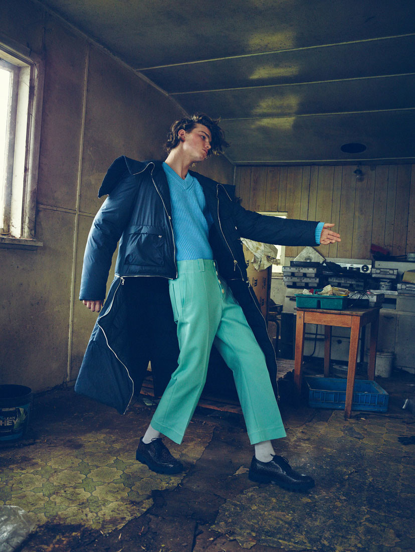 Young man in blue top and teal pants, posing inside with window light, wearing a dark blue jacket with white trim and black shoes.