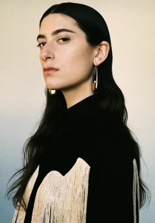 Portrait of a woman with long dark hair wearing a black top with fringed yoke and gold drop earrings against a plain background