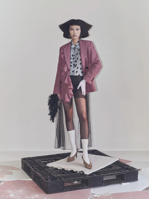 A stylish Asian woman poses in a pink blazer, polka dot top, sheer skirt, patterned heels, and white socks in a fashion studio.