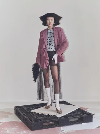 A stylish Asian woman poses in a pink blazer, polka dot top, sheer skirt, patterned heels, and white socks in a fashion studio.