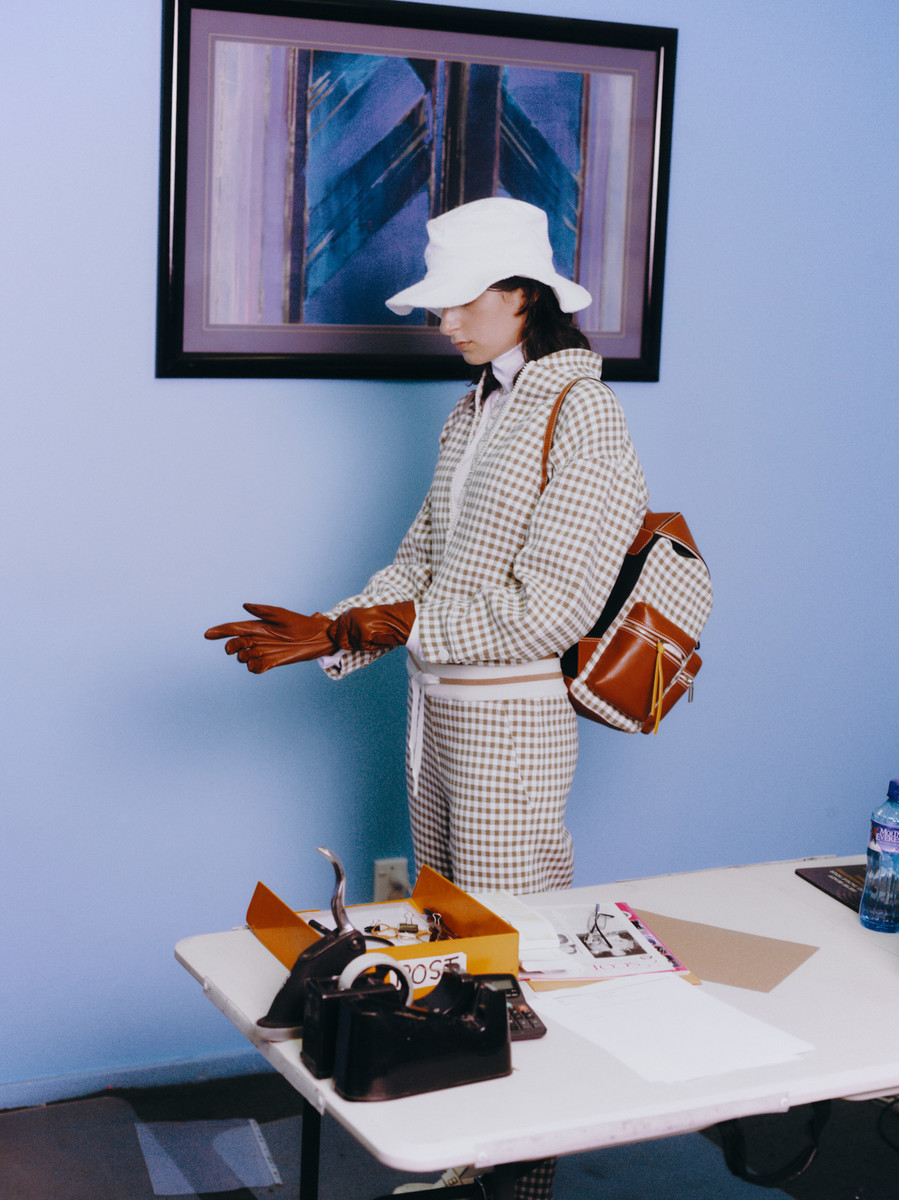 A woman in a patterned suit and hat stands near a table with accessories and a water bottle, with a painting hanging behind her.