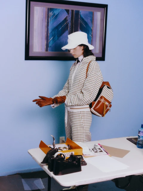 A woman in a patterned suit and hat stands near a table with accessories and a water bottle, with a painting hanging behind her.