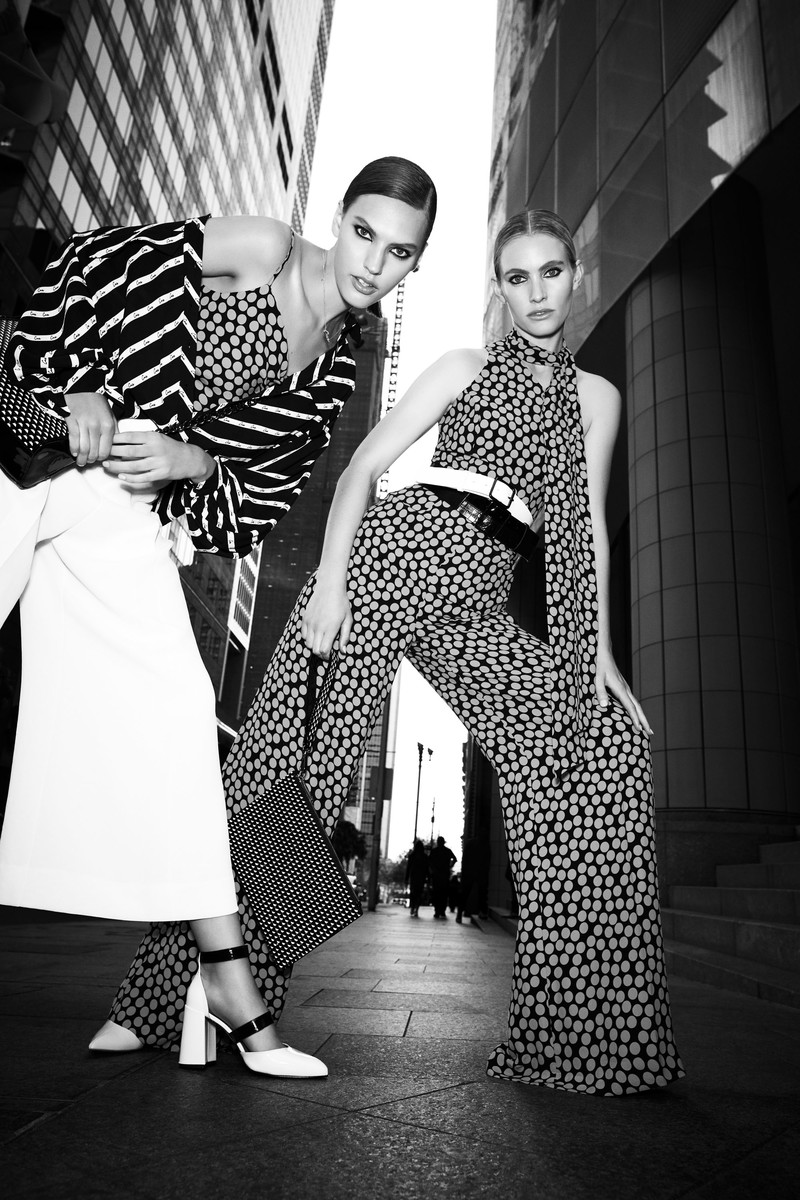 Two fashion models pose in black and white in the city, wearing patterned jumpsuits and dresses, captured at a low angle.