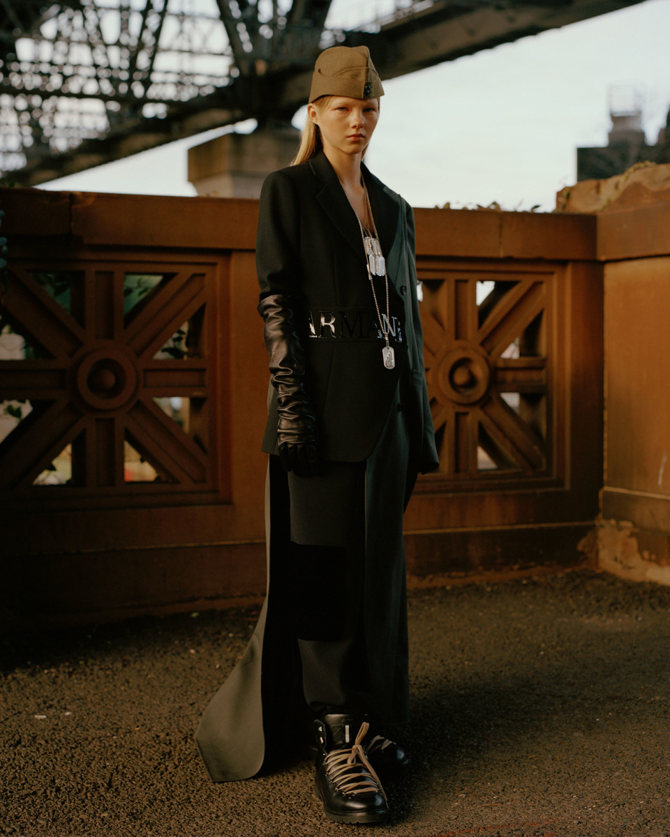 Woman in a long black blazer, pants, and cap poses against a stone ledge with a bridge visible behind her in an outdoor setting.