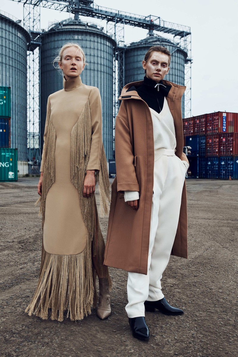 Two fair-skinned fashion models pose outdoors in neutral-toned outfits against an industrial backdrop of silos and shipping containers.