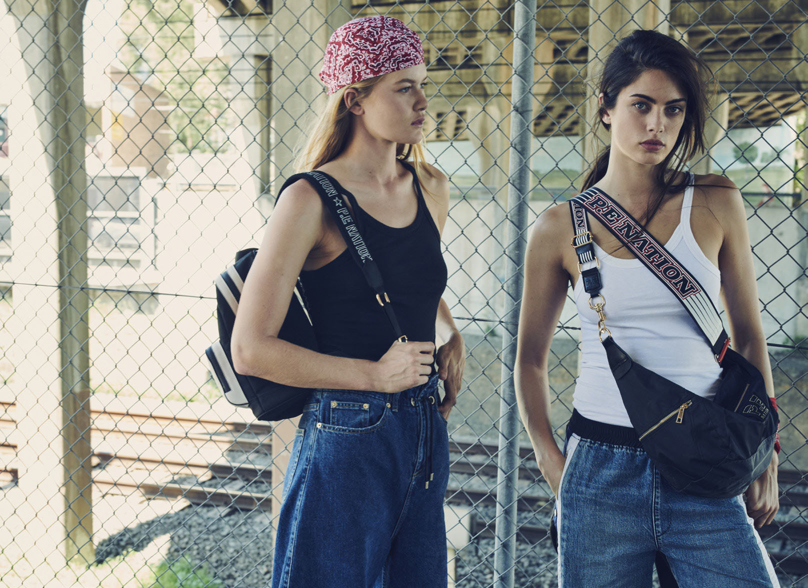 Two women pose outdoors in casual streetwear with shoulder bags, showcasing a fashion-forward urban style against a chain-link fence backdrop.