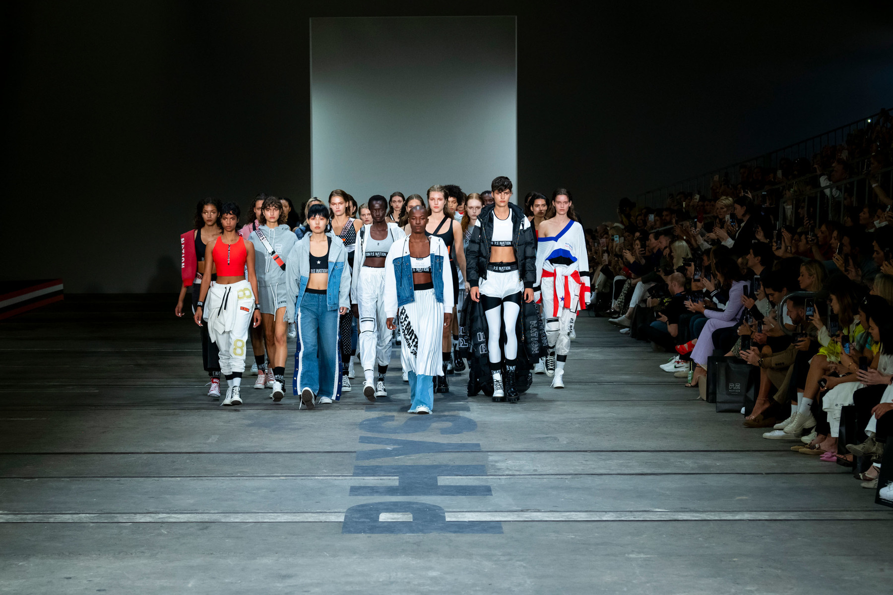 Fashion show on a runway with a group of diverse models showcasing athletic apparel in an indoor venue with audience seating.