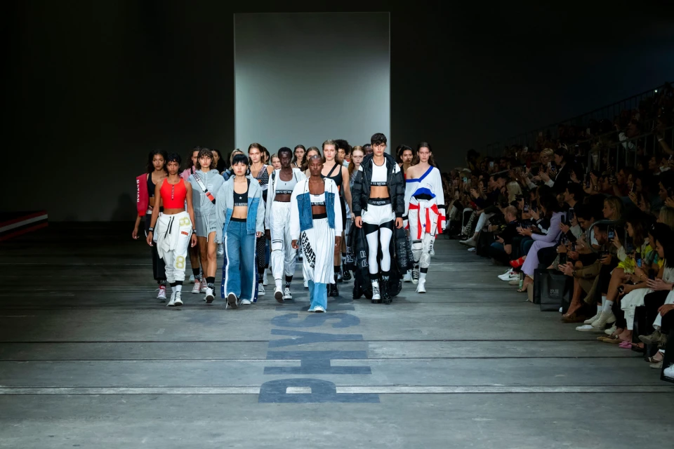 Fashion show on a runway with a group of diverse models showcasing athletic apparel in an indoor venue with audience seating.