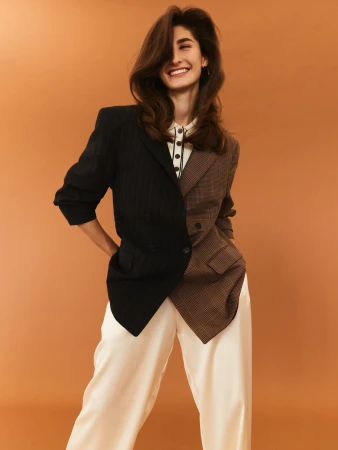 Smiling woman wearing a split-design black and plaid blazer with white trousers against a warm tan background.